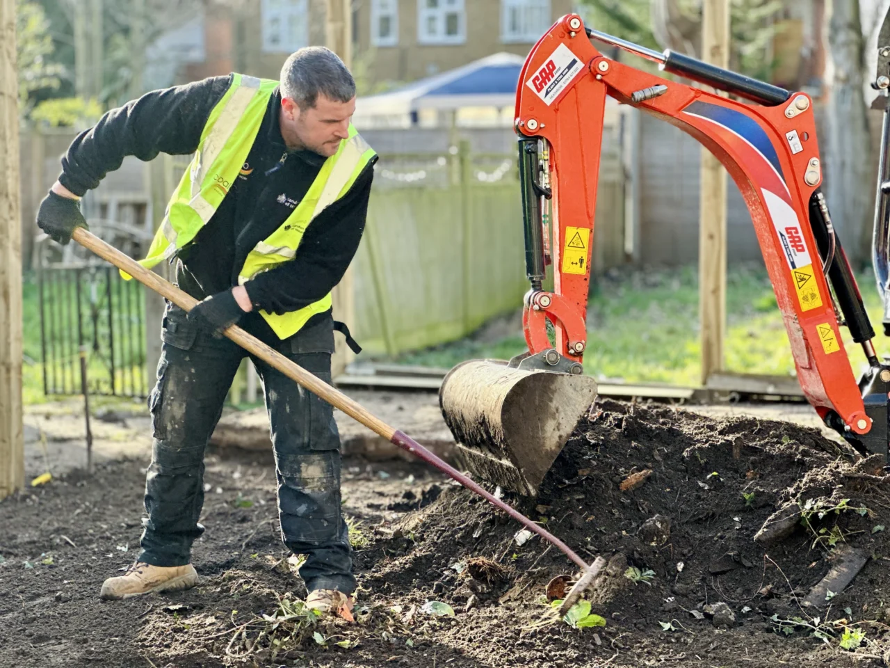 A man in a high visibility jacket, digging a hole with a spade beside a digger